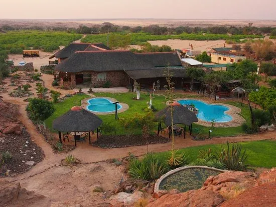 White Lady Lodge from the air at dusk, the thatched main building and pools set against the Ugab riverbed and the desert beyond.