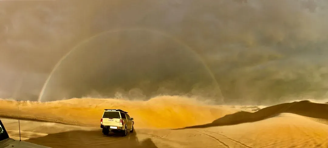 A full rainbow arches over a vehicle crossing the dunes